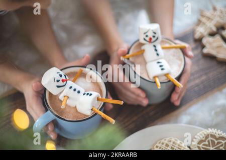 Kleine süße Kinder spielen mit gestrickten Schneemännern, essen Lebkuchen und trinken Kakao mit Marshmallows. Stockfoto