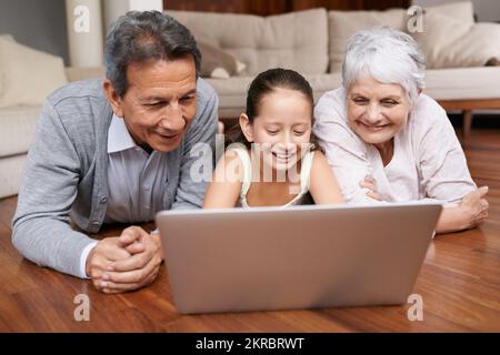 Erstellen eines Social-Media-Profils für Oma und Opa. Ein junges Mädchen und ihre Großeltern liegen auf dem Boden und schauen auf einen Laptop. Stockfoto