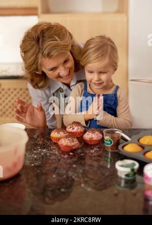 Sie ist stolz auf ihre Cupcake-Kreationen. Ein kleines Mädchen, das Cupcakes mit Hilfe ihrer Großmutter dekoriert. Stockfoto