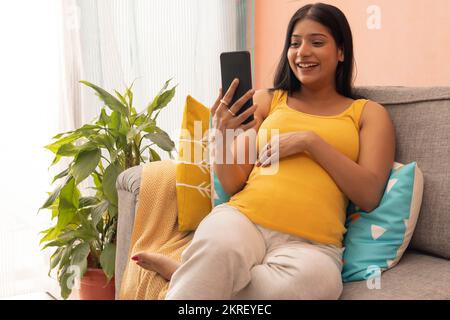 Lächelnde schwangere Frau, die ihr Handy benutzt, während sie sich zu Hause auf dem Sofa entspannt Stockfoto