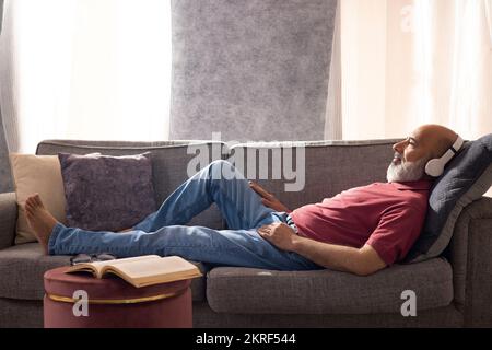 Seniorenmann, der Musik mit Kopfhörern hört, während er zu Hause auf dem Sofa liegt Stockfoto