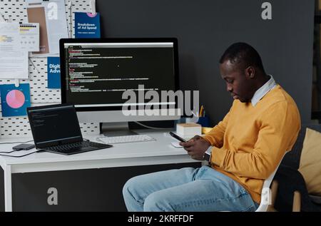 Junger afroamerikanischer Entwickler, der sein Smartphone verwendet, während er am Arbeitsplatz vor dem Computermonitor sitzt und Codes im Büro trägt Stockfoto