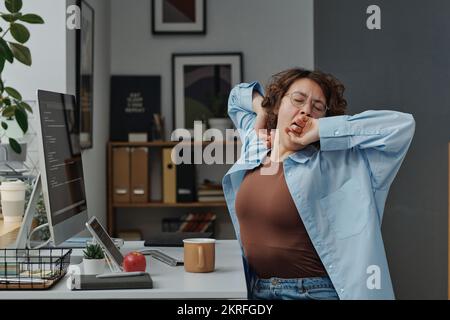 Junge Geschäftsfrau, die es satt hat, am Computer zu sitzen, am Arbeitsplatz zu sitzen und zu gähnen Stockfoto