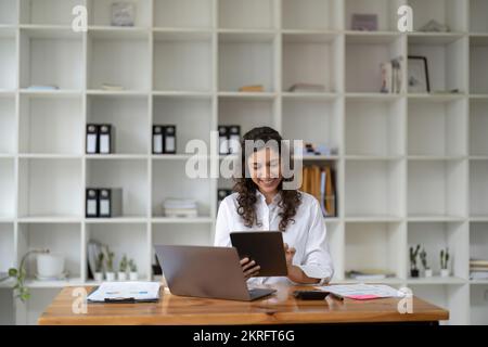 Hübsche, charmante Frau, die sich benimmt, surft, tippt, sucht, Expertisierung, Online-Arbeit auf Computer und Tablet am Arbeitsplatz, Nutzung des wi-Fi-Internets Stockfoto