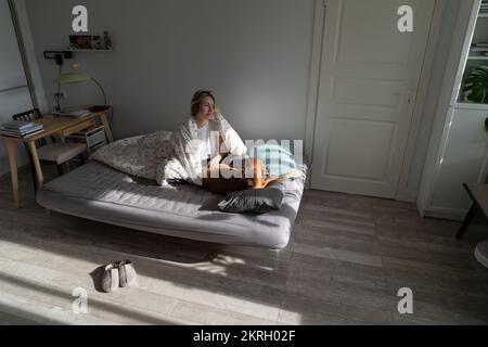 Eine Frau mittleren Alters, die morgens zu Hause mit einem Hund im Bett sitzt und es genießt, allein mit ihrem geliebten Haustier zu leben Stockfoto