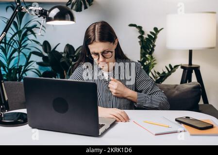 Gestresste junge Frau mit Brille, die einen Herzinfarkt hat, sitzt auf der Couch im Heimbüro. Arbeitskraft erschöpft vom Computer am Arbeitsplatz mit Stress Stockfoto