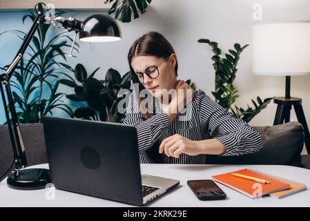 Müde junge brünette Frau mit Brille und schmerzenden Nackenschmerzen arbeitet am Computer, der nachts im Heimbüro auf der Couch sitzt. Nackenschmerzen, eingeklemmte Nerven Stockfoto