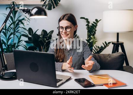 Lächelnde Frau, Videoanrufe für ein Remote-Bewerbungsgespräch mit Laptop, Webcam, Konferenzarbeit, sitzen auf dem Sofa im Heimbüro. Stockfoto