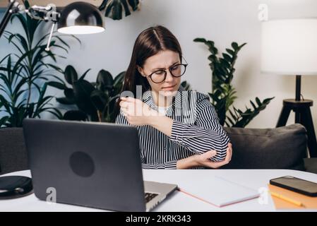 Nahaufnahme einer traurigen jungen Frau mit Brille, die am Arbeitsplatz auf dem Sofa sitzt und Schmerzen und Beschwerden im Ellenbogengelenk erlebt Stockfoto
