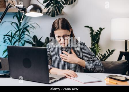 Nahaufnahme einer jungen Frau mit Brille, die am Computer arbeitet, Schwierigkeiten beim Atmen oder Schmerzen in der Brust berührt Brust mit der Hand. Panikattacke, Asthma Stockfoto