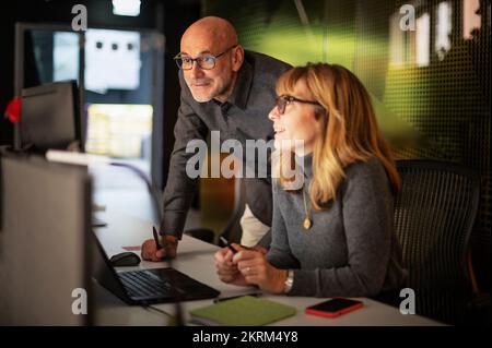 Eine Gruppe ungezwungener Geschäftsleute, die am späten Abend im Büro mit Computern am Tisch sitzen und geschäftliche Informationen analysieren. Teamarbeit. Stockfoto
