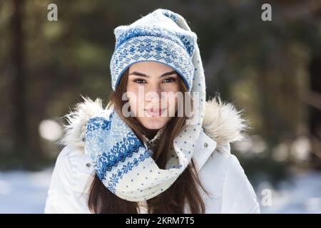 Junge Frau in warmen Outfits, die glücklich in die Kamera schaut, während sie im Winter im sonnigen Wald steht Stockfoto