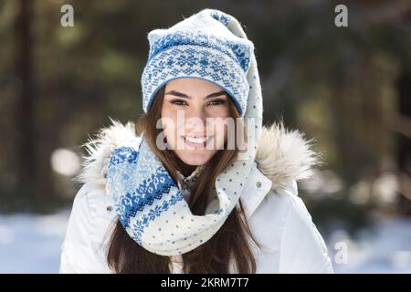 Junge Frau in warmen Outfits, die glücklich in die Kamera schaut, während sie im Winter im sonnigen Wald steht Stockfoto
