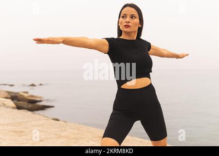 Konzentrierte Sportlerin in enger, schwarzer Sportkleidung, die Yoga an der Küste praktiziert und Virabhadrasana B trainiert, balanciert und meditiert Stockfoto