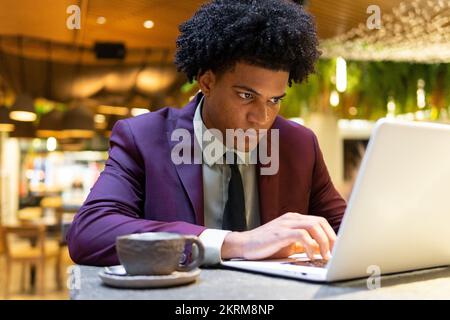 Fokussierter junger afroamerikanischer Freiberufler mit lockigem Haar im stilvollen Anzug, der auf den Bildschirm schaut, während er mit einem Laptop am Tisch mit Kaffee arbeitet Stockfoto