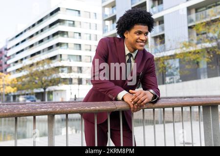 Selbstbewusster junger, eleganter afroamerikanischer Angestellter mit lockigem dunklem Haar in stilvollem Anzug und einer lächelnden Krawatte, die auf einem Fußbett wegsieht Stockfoto