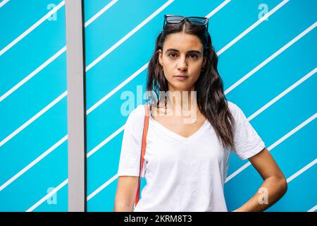 Junge Frau in weißem T-Shirt, die in der Nähe einer bunten Wand mit weißen Streifen auf die Kamera schaut Stockfoto