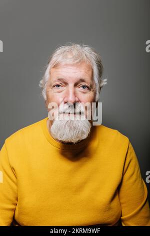 Porträt eines gealterten grauhaarigen Mannes im gelben Pullover, der vor grauem Hintergrund lächelt Stockfoto