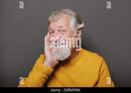 Porträt eines gealterten grauhaarigen Mannes im gelben Pullover, der vor grauem Hintergrund lächelt Stockfoto