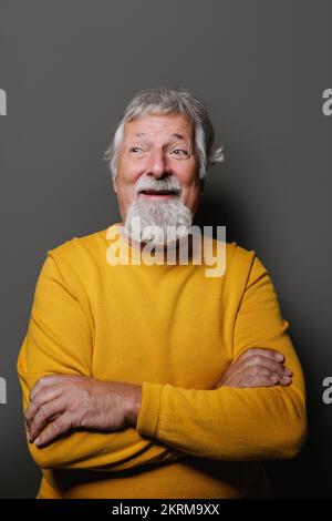 Porträt eines gealterten, grauhaarigen Mannes im gelben Pullover, der vor einem grauen lächelnden Hintergrund wegblickt Stockfoto