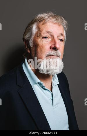 Gereifter, grauhaariger Mann in eleganter Kleidung, der vor grauem Hintergrund steht und wegschaut Stockfoto
