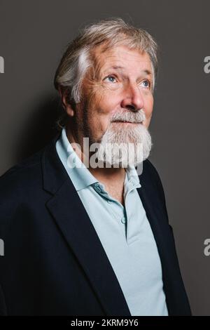 Gereifter, grauhaariger Mann in eleganter Kleidung, der vor grauem Hintergrund steht und nach oben blickt Stockfoto