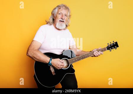 Ein alter Mann mit Bart in lässiger Kleidung spielt Akustikgitarre und singt Lieblingslied, während er in die Kamera schaut Stockfoto