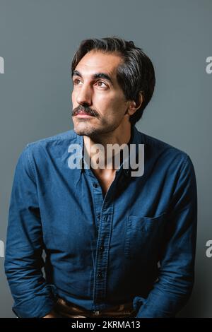 Selbstbewusster junger Mann in blauem Hemd und Jeans, der auf grauem Hintergrund sitzt und wegschaut Stockfoto