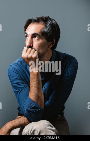Selbstbewusster junger Mann in blauem Hemd und Jeans, der auf grauem Hintergrund sitzt und wegschaut Stockfoto