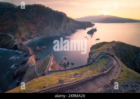 Luftaufnahme einer leeren Straße auf einem Hügel mit grünem Gras in der Nähe des ruhigen Meerwassers während des Sonnenuntergangs in Gaztelugatxe Stockfoto