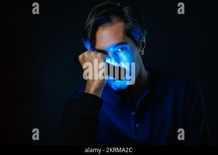 Porträt eines erwachsenen Mannes im lässigen Pullover mit Neonbeleuchtung im Gesicht, der auf die Kamera schaut, während er vor schwarzem Hintergrund steht Stockfoto