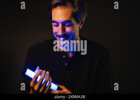 Positiver männlicher Blick nach unten, während er in der Hand glänzende Neonröhre in dunklem Raum vorführt Stockfoto