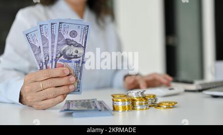 Nahaufnahme: Eine erfolgreiche Frau im asiatischen Alter, die US-Dollar-Scheine, einen Stapel goldener und silberner Münzen und einen Haufen US-Dollar-Scheine auf dem Tisch hält. Stockfoto