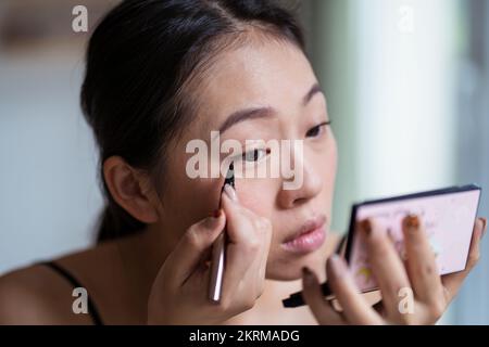 Fokussierte, charmante, ethnische Frau, die beim Make-up Eyeliner auf das Augenlid aufträgt und in den Spiegel schaut Stockfoto