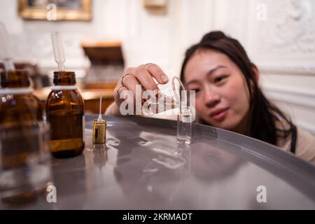 Fokussierter weiblicher Meister, der ätherisches Öl aus dem Glasgefäß in den Kolben auf dem Tisch gießt, während er hausgemachtes Parfüm zubereitet Stockfoto