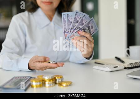 Eine erfolgreiche und wohlhabende asiatische Frau, die US-Dollar-Scheine und -Münzen, einen Haufen Gold- und Silbermünzen und einen Haufen US-Dollar-Scheine auf dem Tisch hält Stockfoto