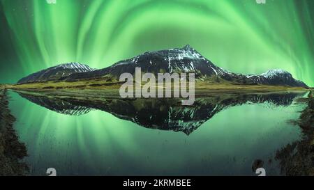 Atemberaubende Aussicht auf schneebedeckte Berge in der Nähe des kristallklaren Sees, der die grün schimmernde Aurora Borealis in Island reflektiert Stockfoto