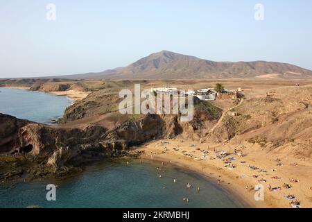 Anonyme Touristen entspannen sich am Sandstrand von Playa de Papagayo, umgeben von felsigen vulkanischen Hügeln an sonnigen Tagen auf Lanzarote Stockfoto