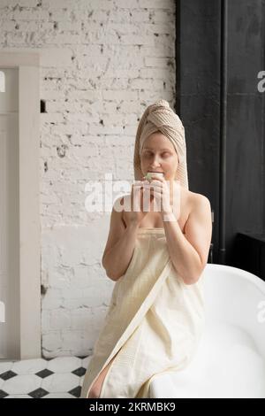Der ganze Körper einer Barfußfrau mit beigefarbenem Handtuch auf dem Kopf entspannt sich im Badezimmer mit weißer Backsteincreme Stockfoto