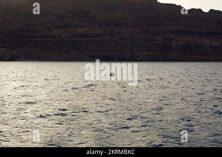 Kleines Motorboot, das im Wellenmeer in der Nähe des riesigen felsigen Vulkans bei Sonnenuntergang in La Graciosa auf den Kanarischen Inseln schwimmt Stockfoto