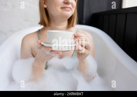 Junge positive Frau mit bob-Haarschnitt im BH, die sich in der Badewanne mit Schaumstoff und Blasen entspannen kann, während sie ein heißes Getränk in einer Tasse mit einer Untertasse genießt, die wegschaut Stockfoto
