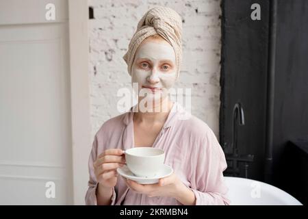 Positive Frau mit Handtuch auf dem Kopf, die neben der Badewanne in einem modernen Badezimmer steht, nachdem sie gebadet hat und weiße Tasse und Untertasse vorführt Stockfoto