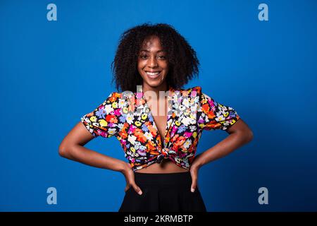 Fröhliche afroamerikanische Frau in stilvollen Kleidern, die mit einem Lächeln auf die Kamera schaut und dabei vor blauem Hintergrund steht Stockfoto
