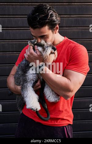 Lächelnder Mann in Sportbekleidung mit dunklen Haaren, der neben einem Metalltor steht und während des Spaziergangs den flauschigen Miniatur-Schnauzer umarmt Stockfoto