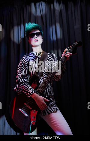 Musikerin in trendigen Outfits und Sonnenbrille, die elektrische Gitarre spielt, während sie vor dunklem Hintergrund steht Stockfoto