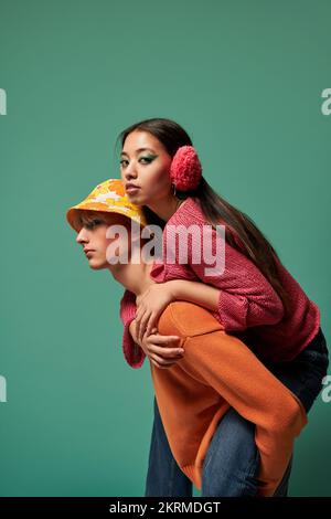 Seitenansicht des Genderqueer-Modells in farbenfroher, stylischer Kleidung mit der jungen philippinischen Frau auf dem Rücken vor türkisfarbenem Hintergrund Stockfoto