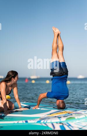 Eine Frau beobachtet einen Mann, der Yoga auf dem Paddleboard macht, während sie Sirasana auf dem Seebad unter wolkenlosem blauen Himmel praktiziert Stockfoto