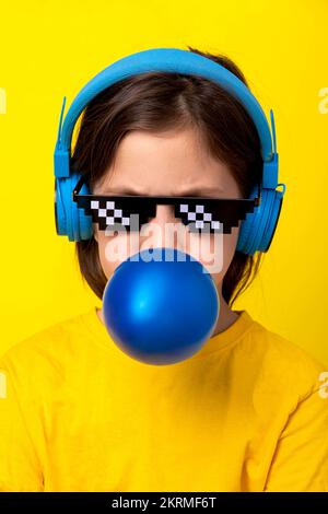 Ein Kind mit braunen Haaren in Kopfhörern hört Musik und bläst einen blauen Kaugummiballon, während es auf gelbem Hintergrund steht Stockfoto