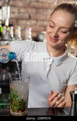 Junge, wunderschöne Barkeeperin gibt Limonade zu einem Cocktailglas. Limonade ist ein Teil vieler Cocktails. Stockfoto