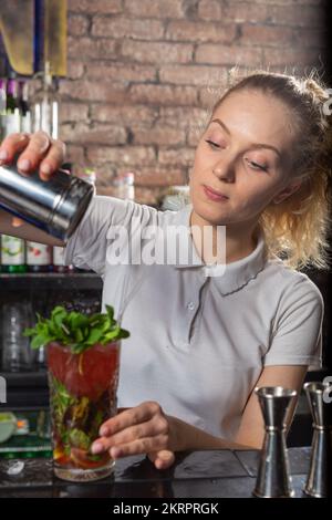 Junge, wunderschöne Barkeeperin streut den Cocktail mit Puderzucker. Die genaue Einhaltung des Rezepts bei der Zubereitung von Cocktails ist der Schlüssel zu s Stockfoto